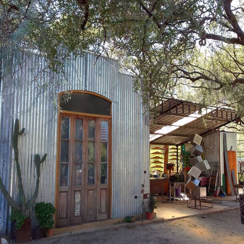 A rustic metal building with wooden doors stands beneath tree branches. An art studio or workshop, visible inside, displays creative sculptures, tools, and colorful materials.