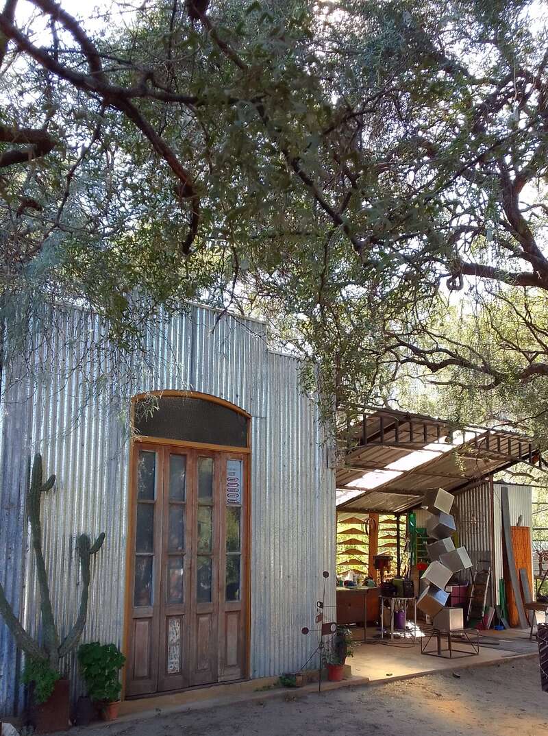 Um prédio rústico de metal com portas de madeira fica embaixo de galhos de árvores. Um estúdio ou oficina de arte, visível em seu interior, exibe esculturas criativas, ferramentas e materiais coloridos.