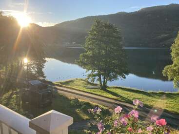 Eine friedliche Seeszene bei Sonnenaufgang, mit Sonnenlicht, das über grüne Hügel fällt, einer ruhigen Spiegelung auf dem Wasser, blühenden Blumen und einem Van, der an einem Baum parkt.