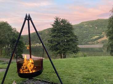 Ein Feuer brennt in einer hängenden Feuerstelle aus Metall auf grünem Gras, mit Blick auf einen friedlichen See mit umliegenden Bäumen und Hügeln unter einem pastellfarbenen Himmel bei Sonnenuntergang.