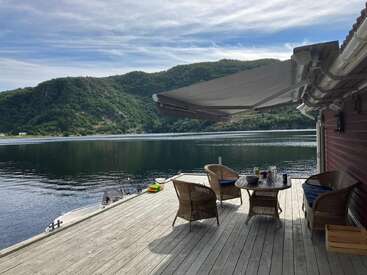 Eine friedliche Seeterrasse mit Korbstühlen und einem Tisch unter einem Sonnenschirm, mit Blick auf ruhige Gewässer und üppig grüne Hügel unter einem teilweise bewölkten Himmel.