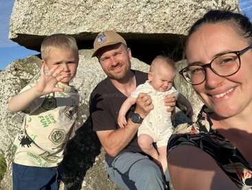 Eine lächelnde Familie posiert im Freien auf felsigem Gelände. Die Mutter trägt eine Brille, der Vater hält ein Baby und ein kleiner Junge winkt fröhlich im hellen Sonnenlicht.