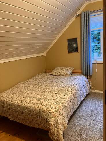 Dieses gemütliche Schlafzimmer hat eine schräge weiße Decke, beige Wände, eine geblümte Tagesdecke, ein kleines Fenster mit blauen Vorhängen, eine Nachttischlampe und ein gerahmtes Bild.