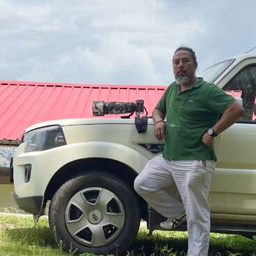 A man in a green shirt and white pants leans against a white SUV. A camera with a long lens rests on the vehicle’s hood.