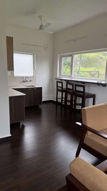 A small, modern kitchen with dark wooden floors, bar seating by a large window, beige armchair, ceiling fan, and bright natural light streaming in.