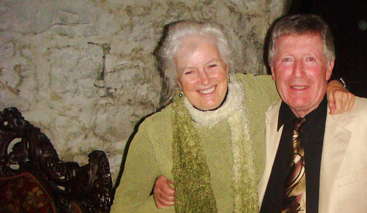 The image shows two older individuals posing for a photo, with the person on the left wearing a light-colored shirt and the person on the right wearing a dark-colored shirt. The background is out of focus.