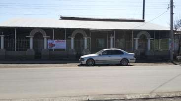 A imagem mostra um carro branco estacionado em frente a um prédio com telhado de metal e janelas em arco, com uma placa onde se lê \"Smart\" em letras vermelhas.