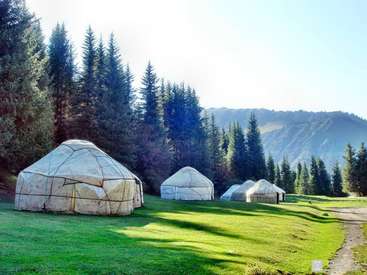 A imagem retrata um campo gramado sereno com quatro yurts, cercado por árvores e montanhas, evocando uma sensação de tranquilidade e beleza natural.