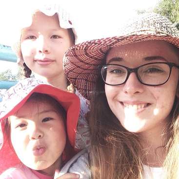 A imagem mostra uma selfie de uma mulher e duas crianças, ambas usando chapéus, posando para a câmera em um ambiente externo ensolarado.