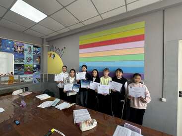 Seven people stand in a classroom, proudly holding certificates. Behind them is colorful wall art and student paintings. Papers and bags are on the table.