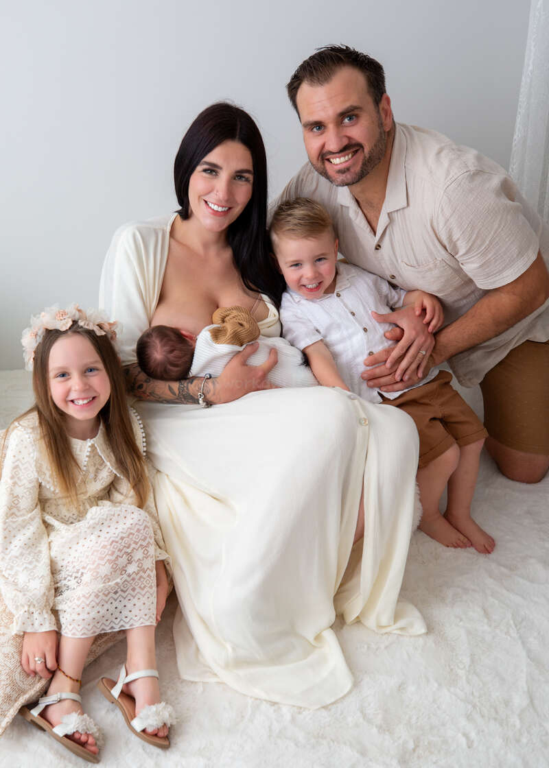 A happy family of five smiles together. The mother breastfeeds a newborn. A young girl, a little boy, and the father gather closely, radiating joy.