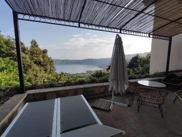 Um terraço coberto com mesas e cadeiras tem vista para uma vegetação exuberante e um lago calmo. O céu está parcialmente nublado, criando uma atmosfera relaxante, cênica e pacífica.