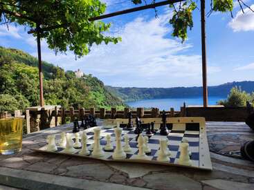 Um tabuleiro de xadrez está em uma mesa rústica com um copo de bebida, com vista para um lago sereno e colinas verdejantes sob um céu claro e parcialmente nublado.