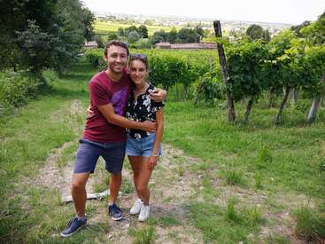 Um casal sorridente posa junto ao ar livre em um caminho de vinhedos, cercado por videiras verdejantes e uma paisagem campestre com árvores e campos distantes ao fundo.