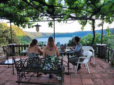 Três mulheres desfrutam de uma refeição ao ar livre sob uma pérgula frondosa, com vista para um lago sereno cercado por colinas exuberantes, criando uma atmosfera pacífica e cênica.