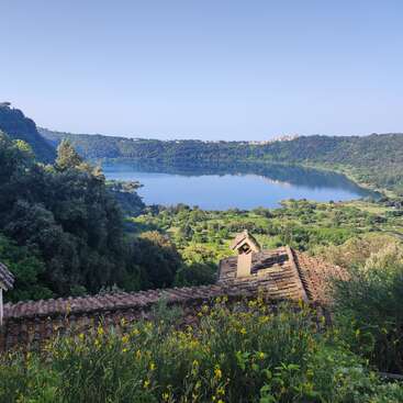 A serene lake is surrounded by lush green hills and trees. Yellow wildflowers and rustic rooftops are in the foreground under a clear blue sky.
