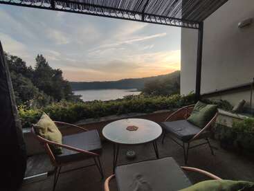 Um pátio aconchegante com duas cadeiras, uma mesa e almofadas tem vista para um lago sereno e uma vegetação exuberante ao pôr do sol, criando uma atmosfera tranquila e convidativa.