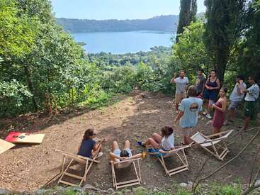 Um grupo de amigos relaxa ao ar livre em espreguiçadeiras com vista para um lago panorâmico, cercado por árvores, desfrutando de conversas e da natureza em um dia ensolarado.