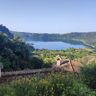 A serene lake is surrounded by lush green hills and trees. Yellow wildflowers and rustic rooftops are in the foreground under a clear blue sky.