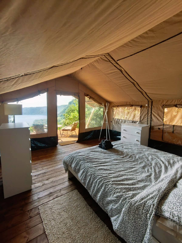 This image shows a cozy glamping tent with a comfortable bed, wooden floor, white furniture, and large windows overlooking beautiful hills and an outdoor seating area.