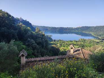 Un lago sereno está rodeado de frondosas colinas verdes y árboles. En primer plano, flores silvestres amarillas y tejados rústicos bajo un cielo azul despejado.