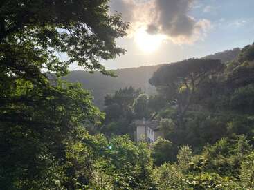 The image depicts a serene scene of a house nestled among lush greenery and trees, with a majestic mountain range in the background under a cloudy sky.