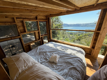 This cozy wooden cabin bedroom features large windows overlooking a scenic lake and lush green hills, offering natural light, serenity, and breathtaking nature views from bed.