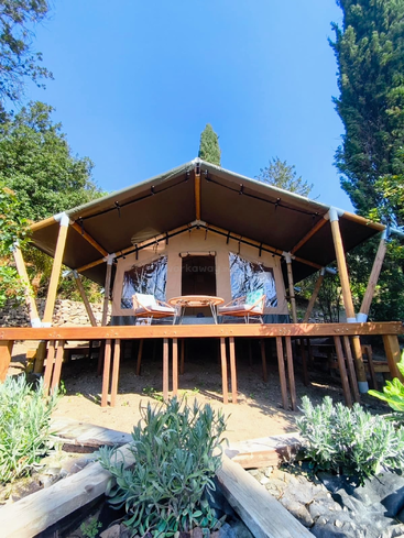 This image shows a glamping tent on a raised wooden deck, with two chairs and a table outside, surrounded by greenery under a clear blue sky.