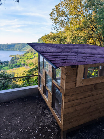 A small wooden shed with glass windows stands on a dirt patch, overlooking a lush green valley and serene lake, surrounded by trees under a blue sky.