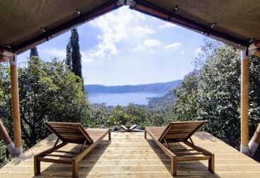 Two wooden lounge chairs sit on a wooden deck under a tent roof, overlooking a lush green forest and a serene lake in the distance.