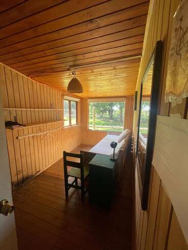 Dieses gemütliche Holzzimmer verfügt über ein Einzelbett, einen kleinen grünen Schreibtisch, einen einfachen Stuhl, große Fenster mit Blick auf die Landschaft, eine Hängelampe und eine minimalistische, rustikale Einrichtung.