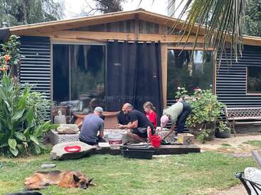 Cuatro personas trabajan juntas en un proyecto frente a una cabaña moderna. Están rodeadas de una exuberante vegetación. Un pastor alemán descansa sobre la hierba cerca de allí.