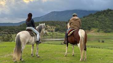 Dos personas montan a caballo frente a un paisaje verde y tranquilo, con colinas onduladas y un lago, bajo un cielo nublado con montañas al fondo. Ambiente apacible.