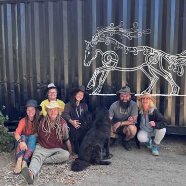Six personnes souriantes, coiffées de chapeaux, sont assises ensemble à l'extérieur, devant un mur noir orné d'un dessin artistique représentant un cheval blanc, en compagnie de deux grands chiens noirs ; elles ont l'air heureuses et détendues.