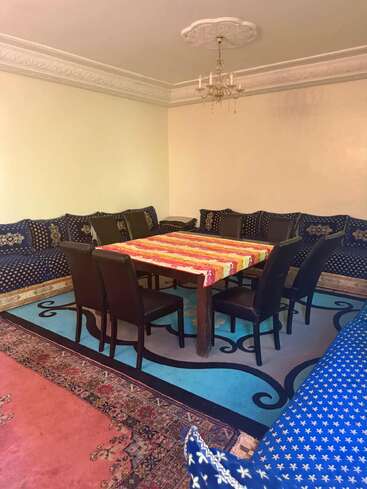 Cette image montre une salle à manger traditionnelle avec une nappe colorée, entourée de chaises noires et de canapés à motifs bleus. Le plafond est orné de moulures et d'un lustre.