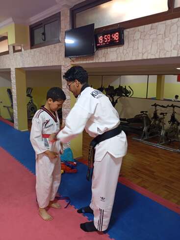 Un instructeur d'arts martiaux aide un jeune garçon à attacher sa ceinture dans un gymnase équipé d'appareils d'exercice, sous une horloge numérique indiquant 19:59:48.