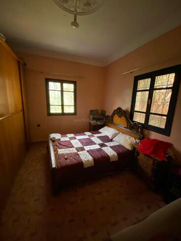 Une chambre confortable avec des murs de couleur pêche, deux fenêtres, une armoire en bois, un sol à motifs, un grand lit avec un édredon à carreaux rouges et une table de nuit avec des cadres.