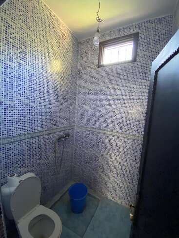 Cette image montre une petite salle de bains avec des murs carrelés de mosaïque bleue, des toilettes, un bidet à main, un seau bleu, du papier hygiénique, une fenêtre au-dessus et une ampoule suspendue apparente.