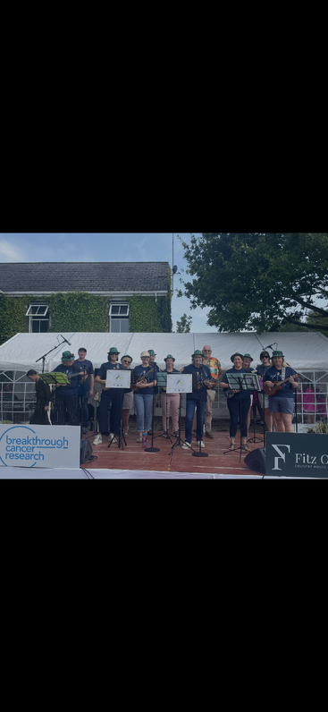 A cheerful group poses with musical instruments on an outdoor stage at a charity event, supporting Breakthrough Cancer Research. Bright hats, banners, and smiles create a lively atmosphere.