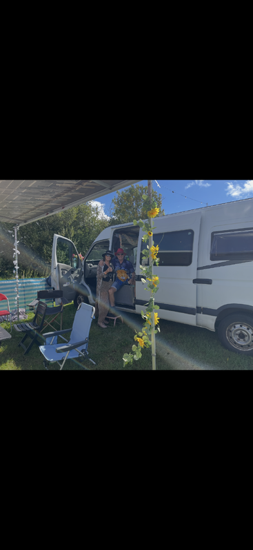 A couple relaxes by their white van under a canopy decorated with sunflowers. Camping chairs, blue sky, and greenery create a cheerful outdoor atmosphere.