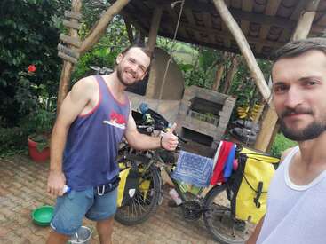 Zwei Männer posieren fröhlich draußen, einer zeigt den Daumen nach oben. In der Nähe hängen Fahrräder mit gelben Gepäcktaschen und Wäsche. Ein rustikaler Holzunterstand und üppiges Grün vervollständigen die Szene.