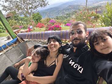 Eine vierköpfige Familie entspannt sich in einer Hängematte im Freien, umgeben von leuchtenden Blumen, üppigem Grün und den Bergen im Hintergrund, und genießt einen fröhlichen und malerischen Moment.