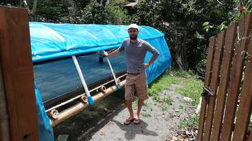 Un homme coiffé d'une casquette se tient à côté d'une longue structure à ossature de bambou recouverte de plastique bleu, à l'extérieur, près d'une clôture en bois et entouré d'arbres et de plantes verts.