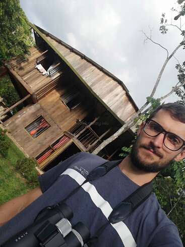 Un homme à lunettes et barbu prend un selfie en plein air, un jumelle à la main. Derrière lui se trouve une maison rustique en bois, entourée de verdure, sous un ciel nuageux.