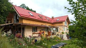 Uma casa grande e rústica com telhado vermelho e várias claraboias fica em meio à vegetação. O jardim é animado, com flores, plantas e uma área de pátio aconchegante.