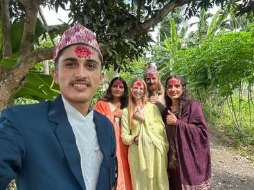 A group of five people, dressed in traditional clothing with tikas on their foreheads, smiling and posing outdoors amidst lush greenery, enjoying a festive occasion.