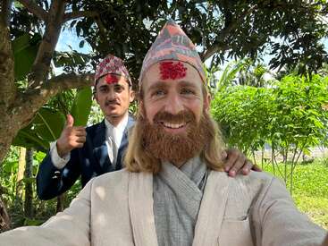 Two men wearing traditional Nepali attire and Dhaka topi, with red tika on their foreheads, smile posing outdoors under trees, exuding cultural pride and friendship.