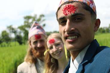 Three people, wearing traditional Nepali hats and tika on their foreheads, stand outdoors in a green field, smiling joyfully under bright sunlight. Celebratory mood.