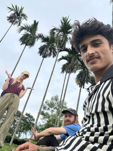 Three people pose outdoors under tall palm trees. A woman stands shrugging, while two men sit on the grass, one taking the selfie. The sky is cloudy.