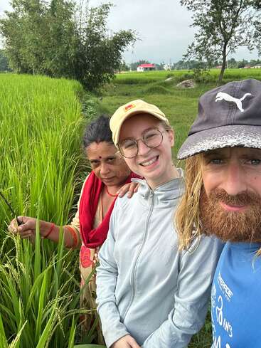 Three people stand together, smiling in a lush green paddy field. One holds rice plants; the background features trees, a red-roofed house, and cloudy sky.